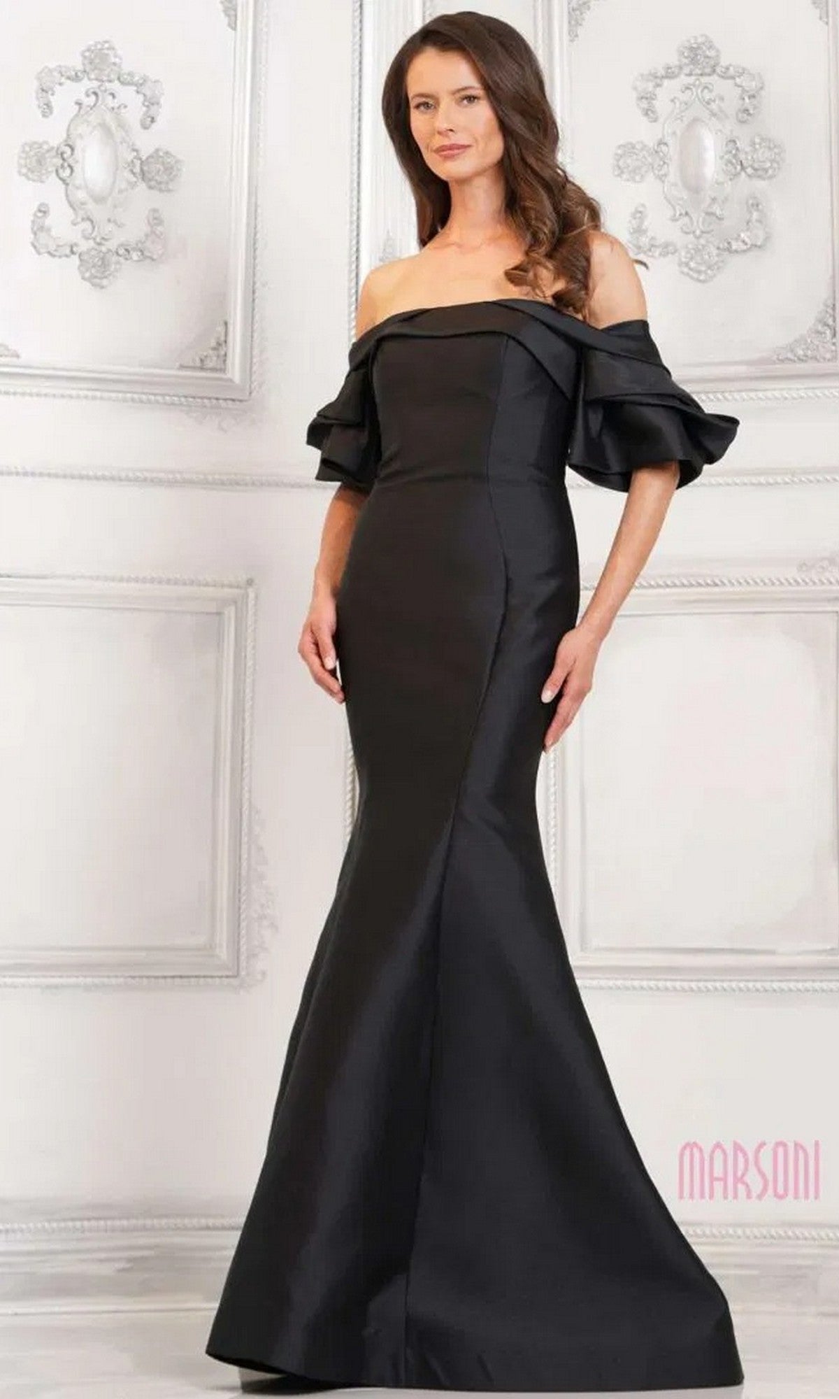 A woman stands in an elegant, white-paneled room wearing the Marsoni MV1290 long formal dress—an off-the-shoulder, floor-length black gown with a mermaid silhouette and ruffled sleeves.